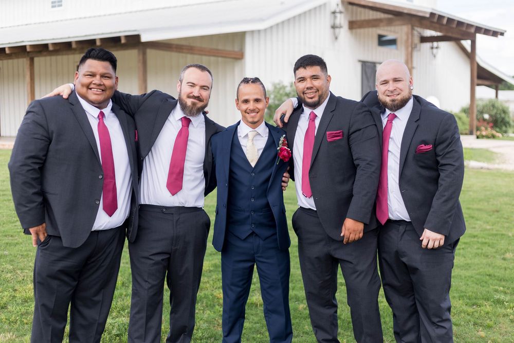 Groom in navy suit poses with four groomsmen in charcoal suits and burgundy ties at outdoor wedding venue.