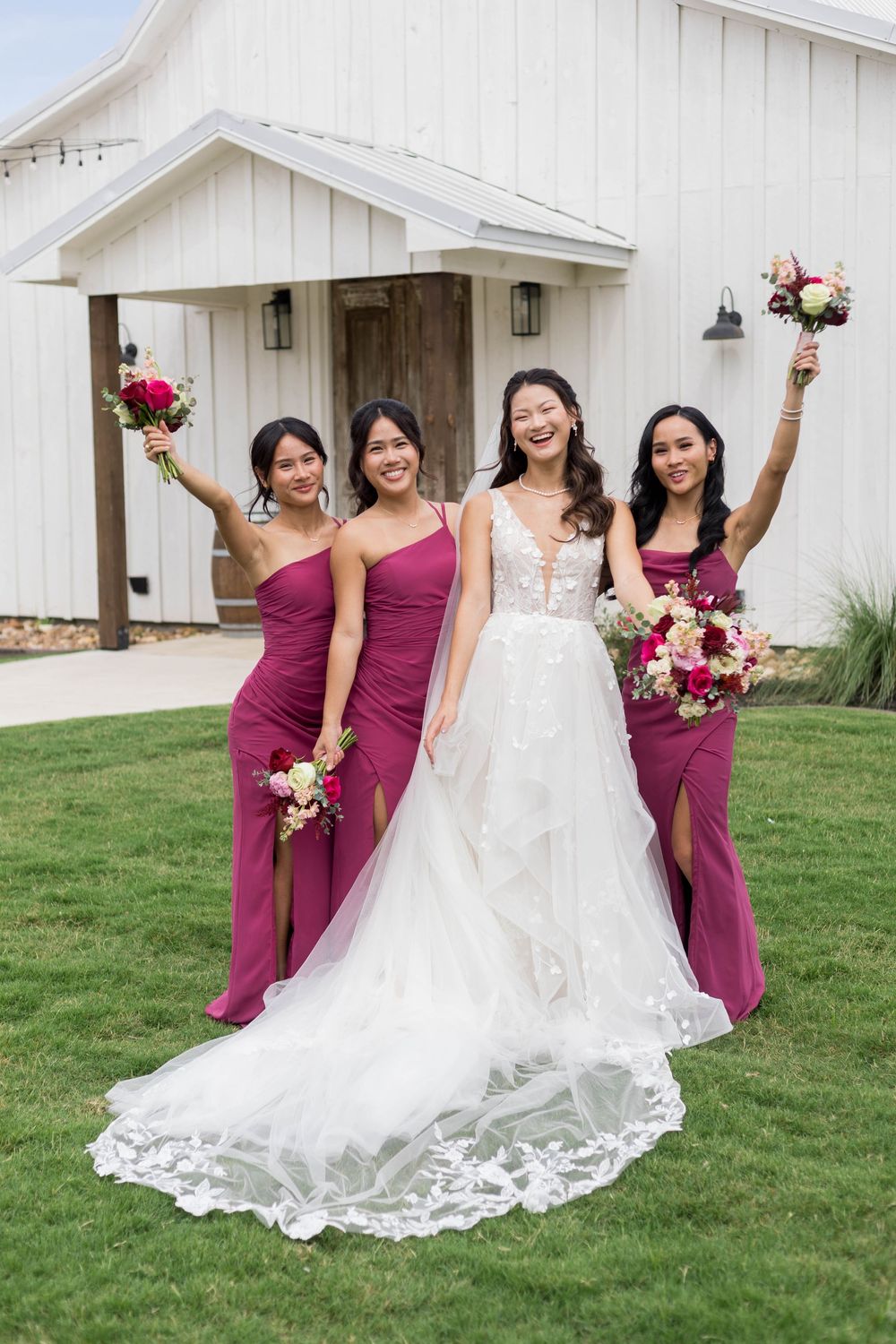 Bride in white lace gown with three bridesmaids in burgundy dresses holding bouquets outdoors at a white barn venue.