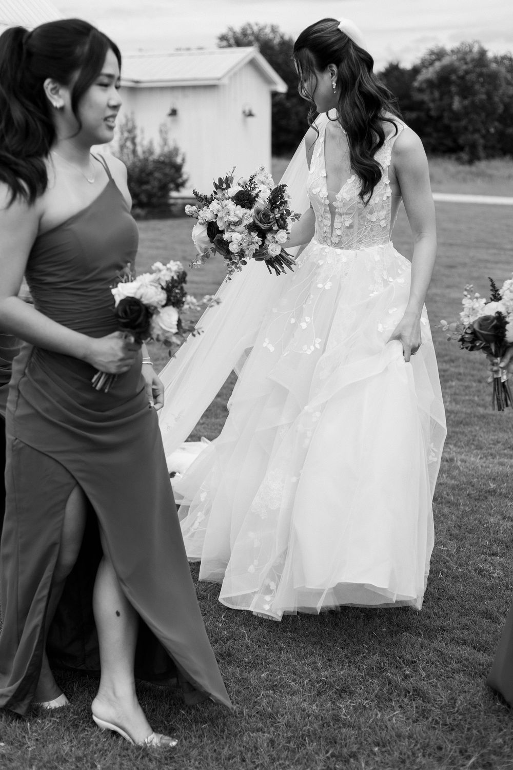 Black and white photo of a bride in a lace ball gown holding a bouquet, walking with a bridesmaid in a gray dress.