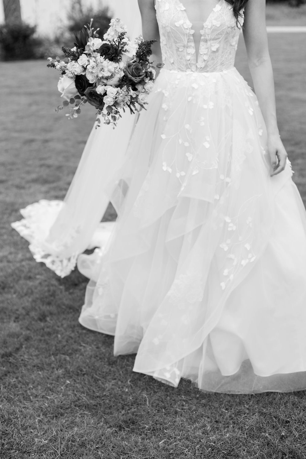 Black and white photo of a bride in a floral lace ballgown holding a bouquet while walking on grass.