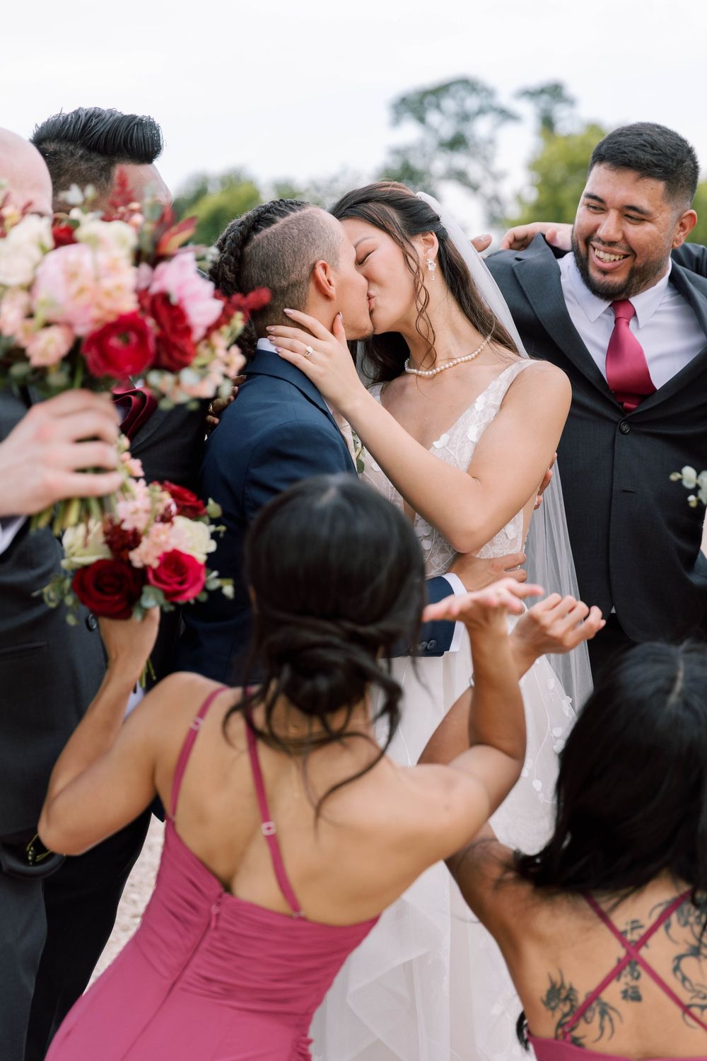 Joyful wedding party surrounds couple sharing first kiss, with red and pink floral bouquets at outdoor ceremony.
