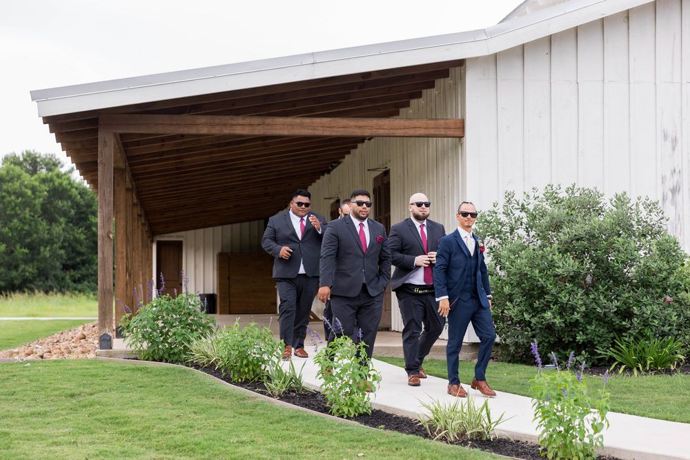 Groomsmen in navy suits and red ties walk confidently outside a modern white barn wedding venue on a sunny day.