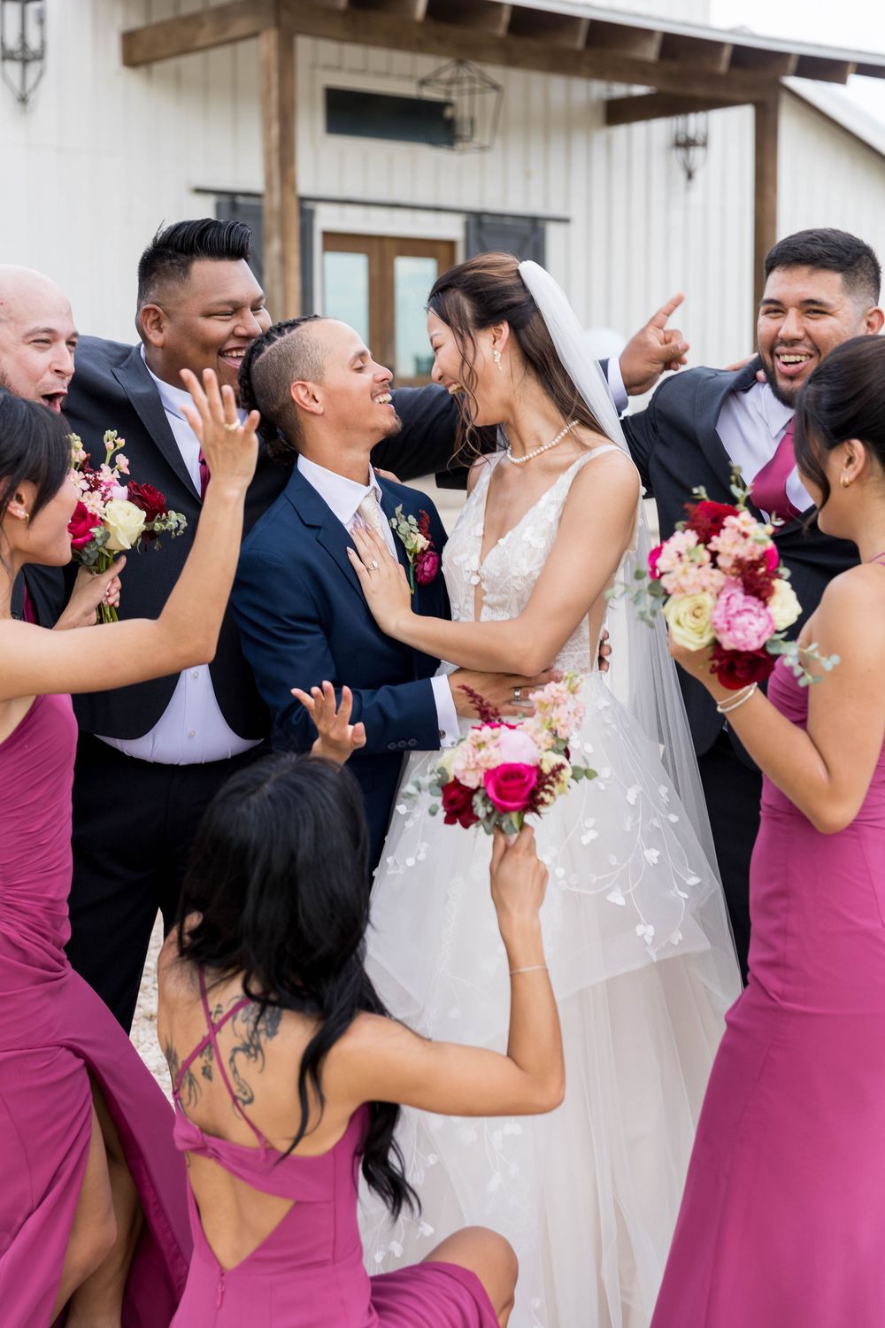 Bride and groom share a romantic moment surrounded by cheering wedding party in magenta dresses and navy suits outdoors.