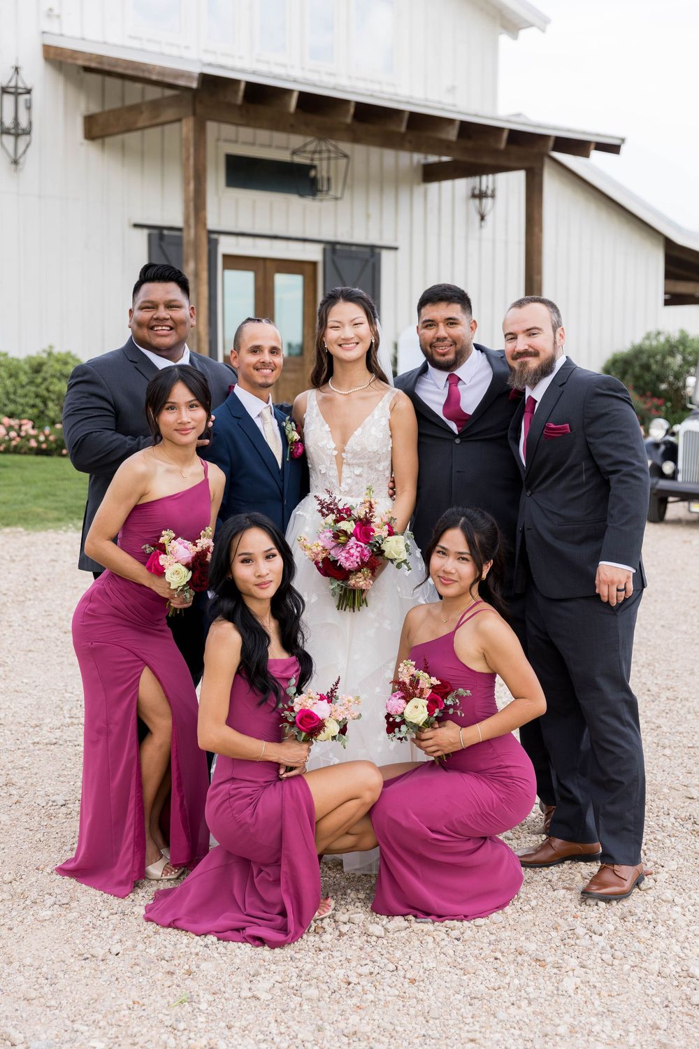Wedding party poses outside barn venue, bride in white gown with bridesmaids in magenta dresses and groomsmen in navy suits.