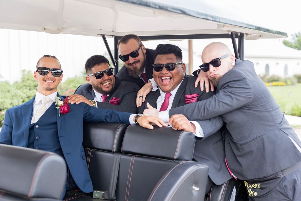 Five groomsmen in gray suits with red boutonnieres pose together smiling in a golf cart at a wedding.