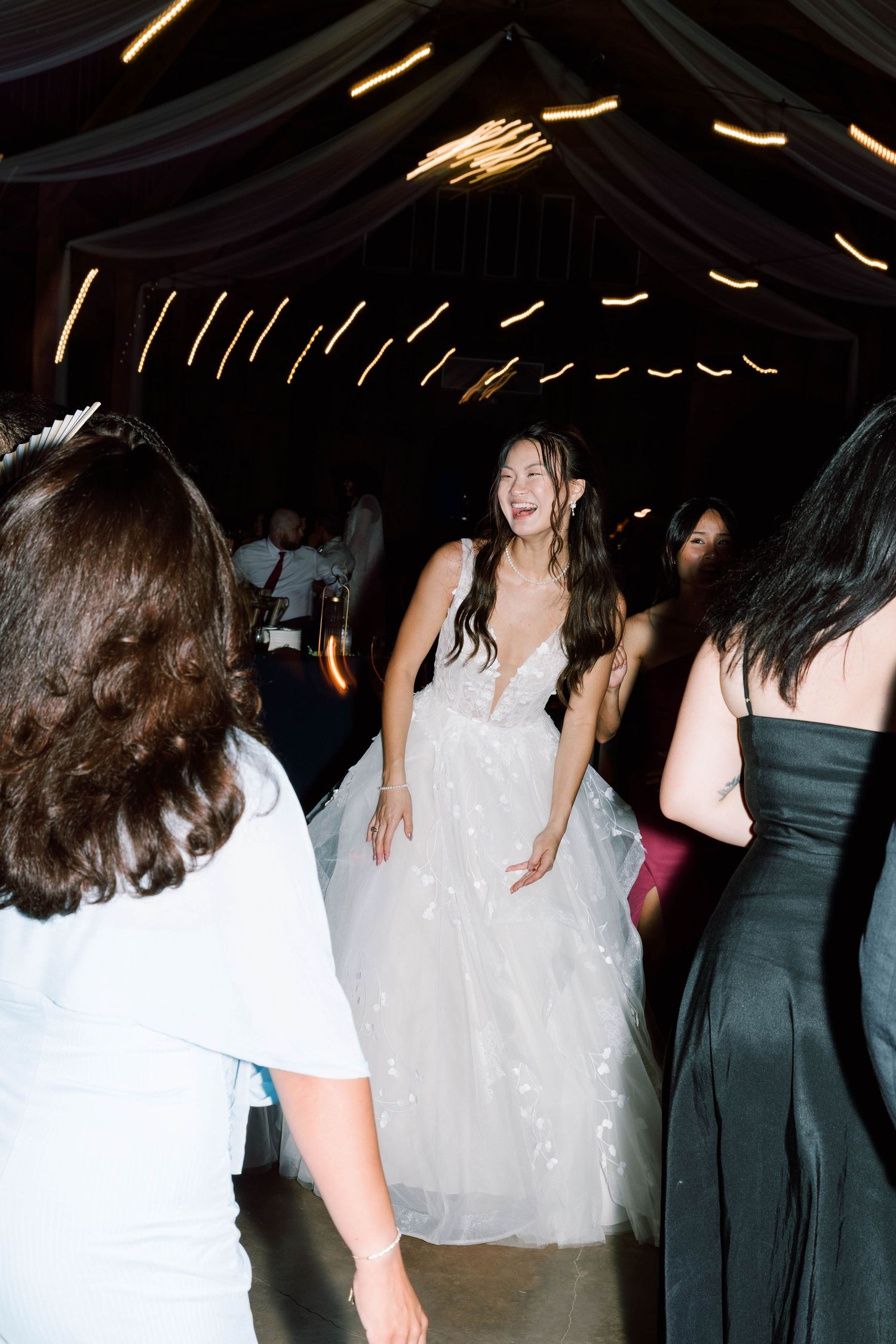 Bride in a white floral tulle gown laughing joyfully on the dance floor surrounded by guests at a wedding reception.