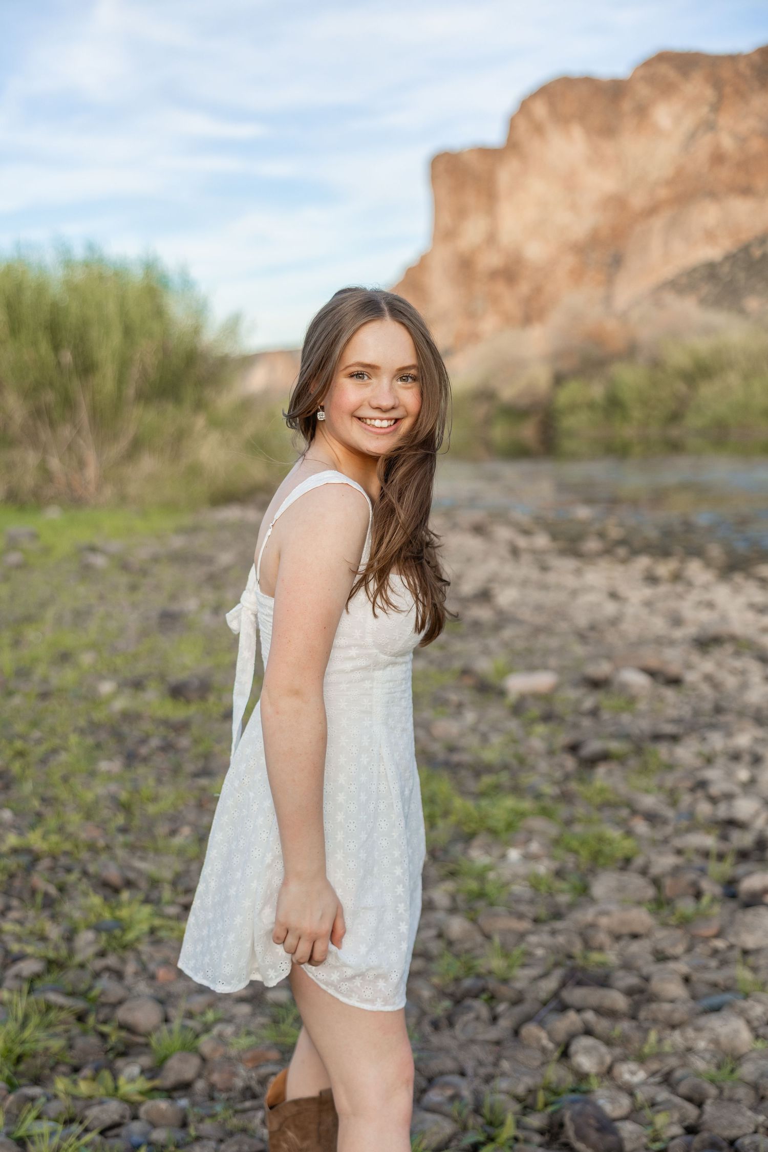 Young woman in white sundress smiling outdoors near rocky riverbed with desert cliffs and green shrubs in background.