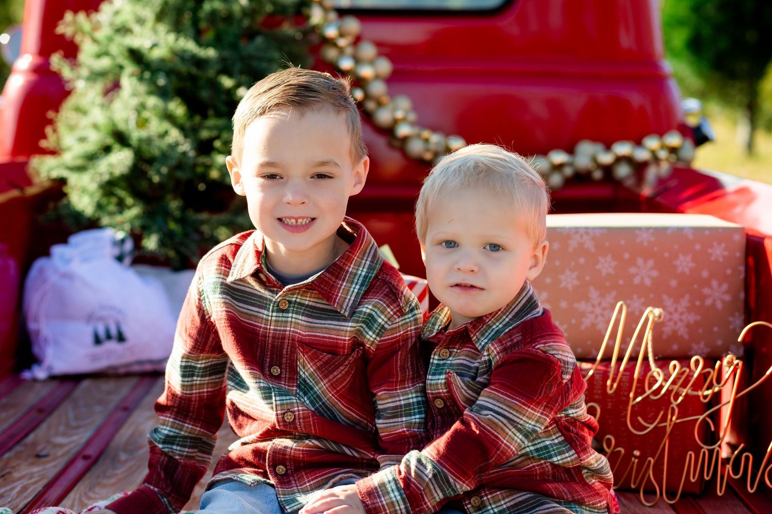Two boys in matching red plaid shirts pose in a red truck bed with Christmas tree and gifts.