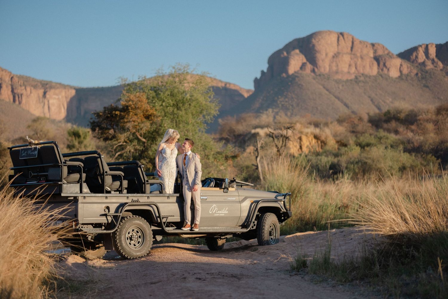 Couple stands by safari jeep in arid landscape with dramatic rocky mountains in background.