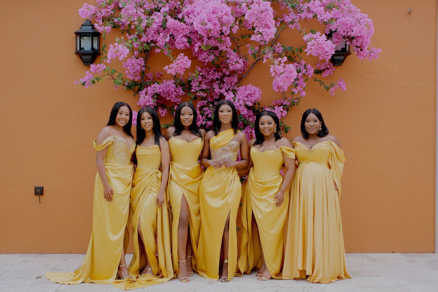 Seven bridesmaids in matching golden yellow satin dresses pose before a vibrant pink bougainvillea wall.