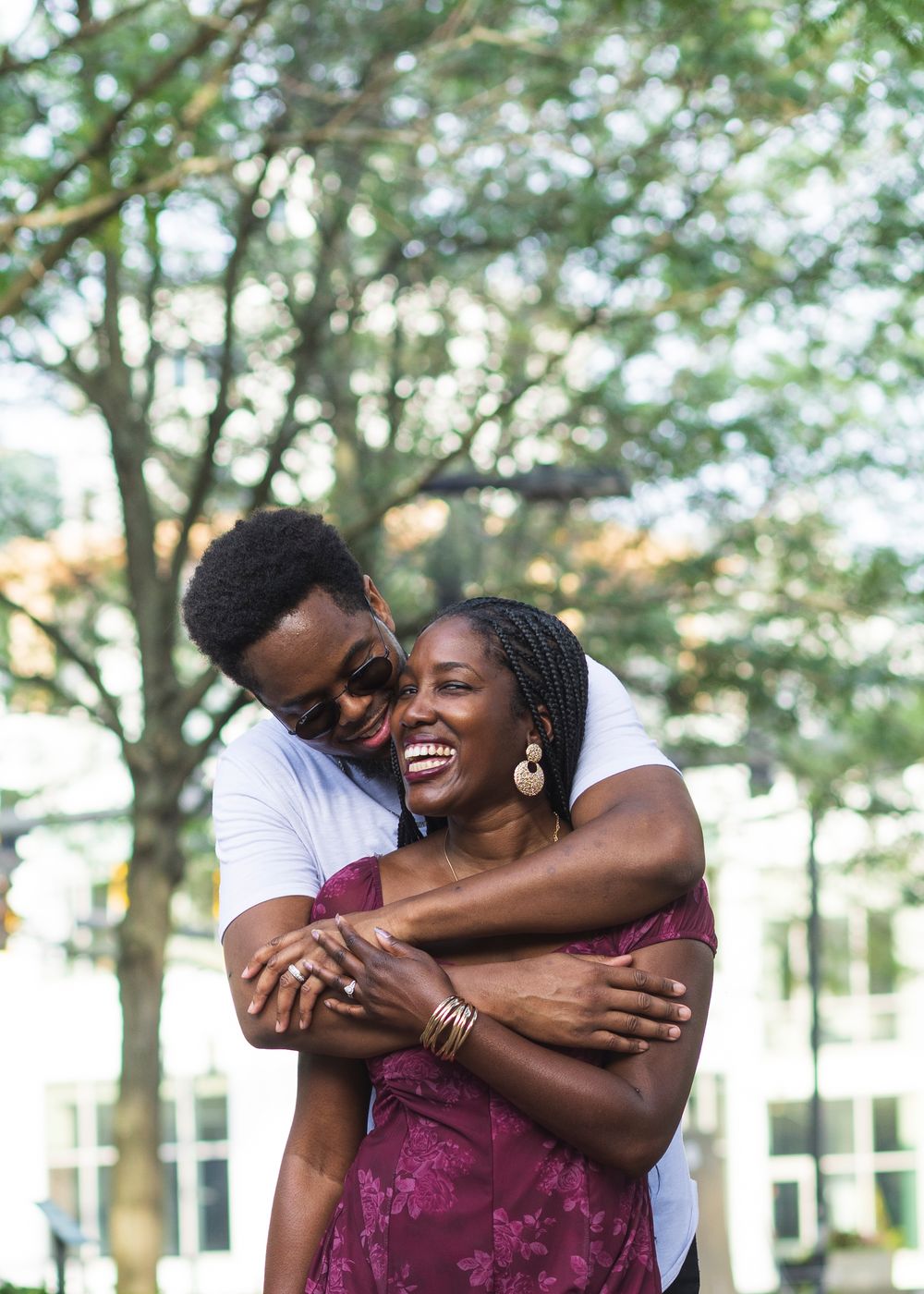 A joyful couple embraces outdoors, the woman laughing warmly as the man hugs her from behind near lush green trees in Lexington, Kentucky.