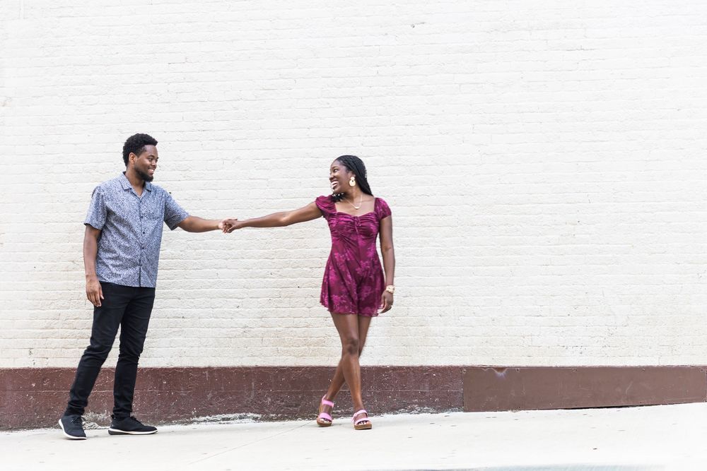 A young couple holds hands at arm's length, smiling at each other against a white brick wall outdoors with Lexington couple photographer