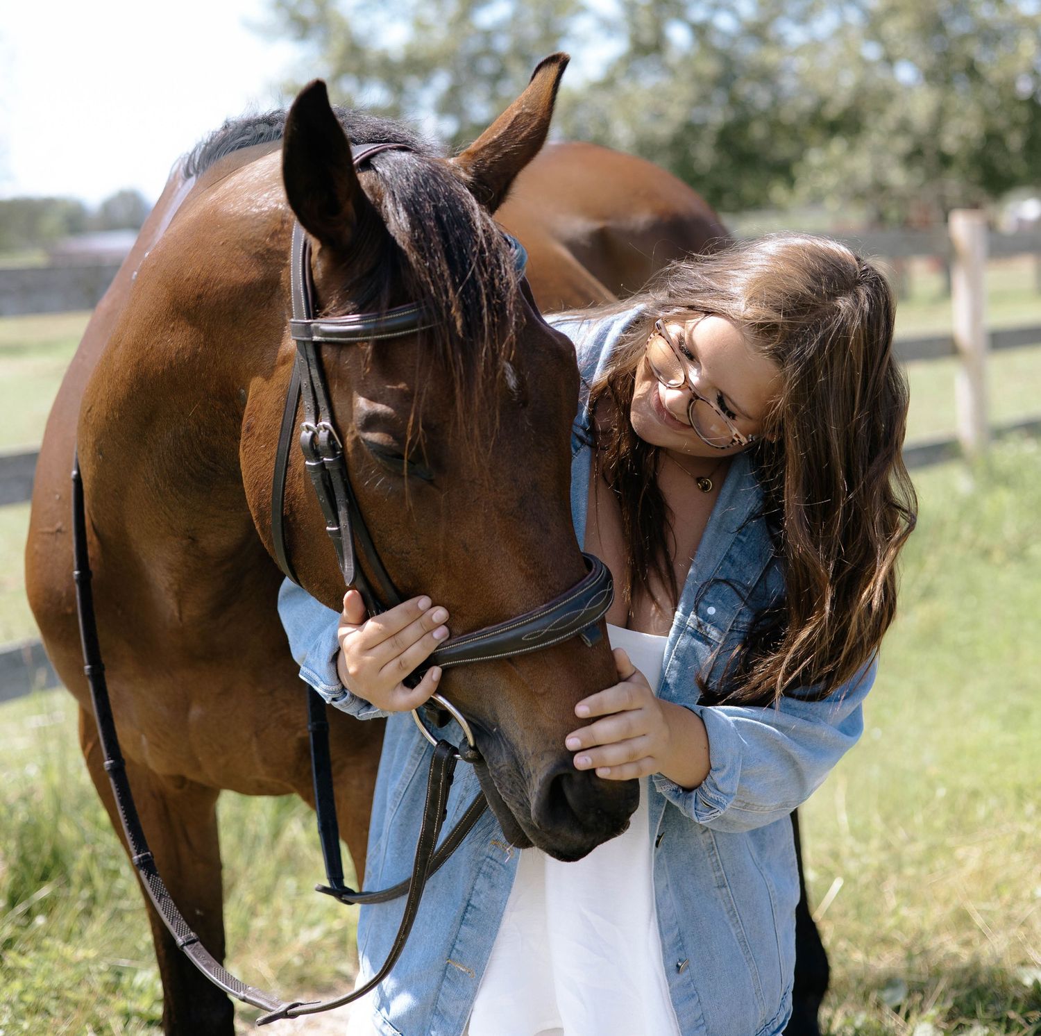 A young woman in a denim jacket affectionately nuzzles a bay horse wearing a bridle in a sunny pasture.