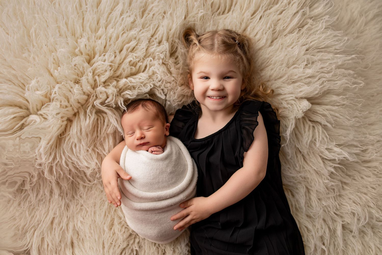 A smiling toddler girl in black dress poses with her swaddled newborn sibling on a fluffy cream rug.