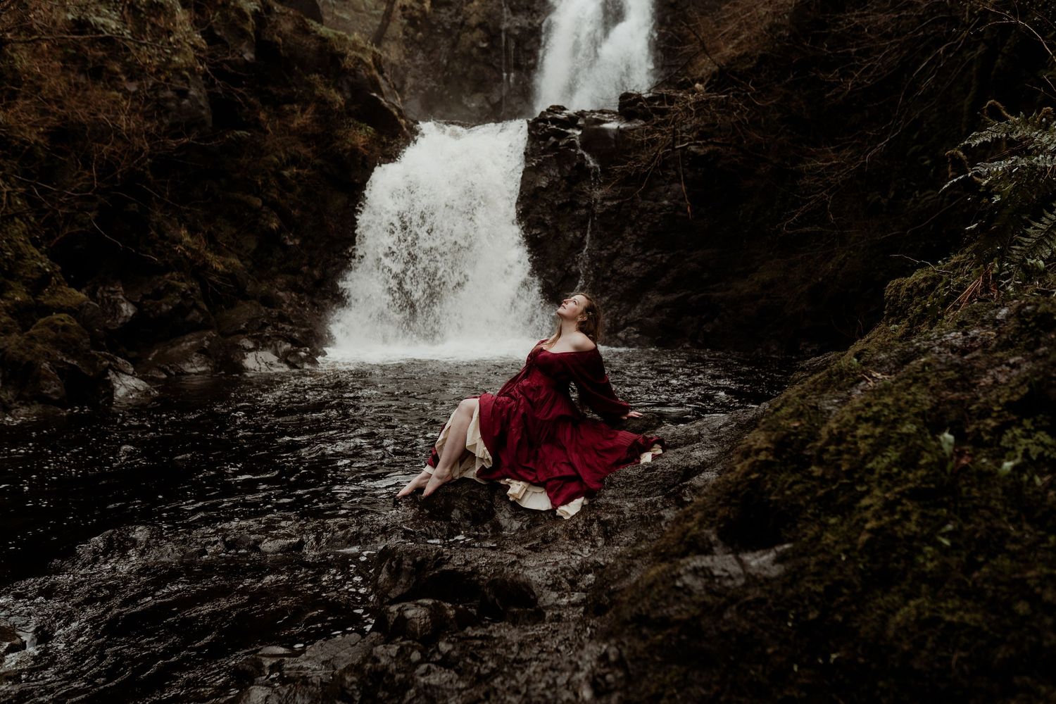 Woman in red dress sitting on rocks beside a dramatic waterfall surrounded by mossy cliffs.