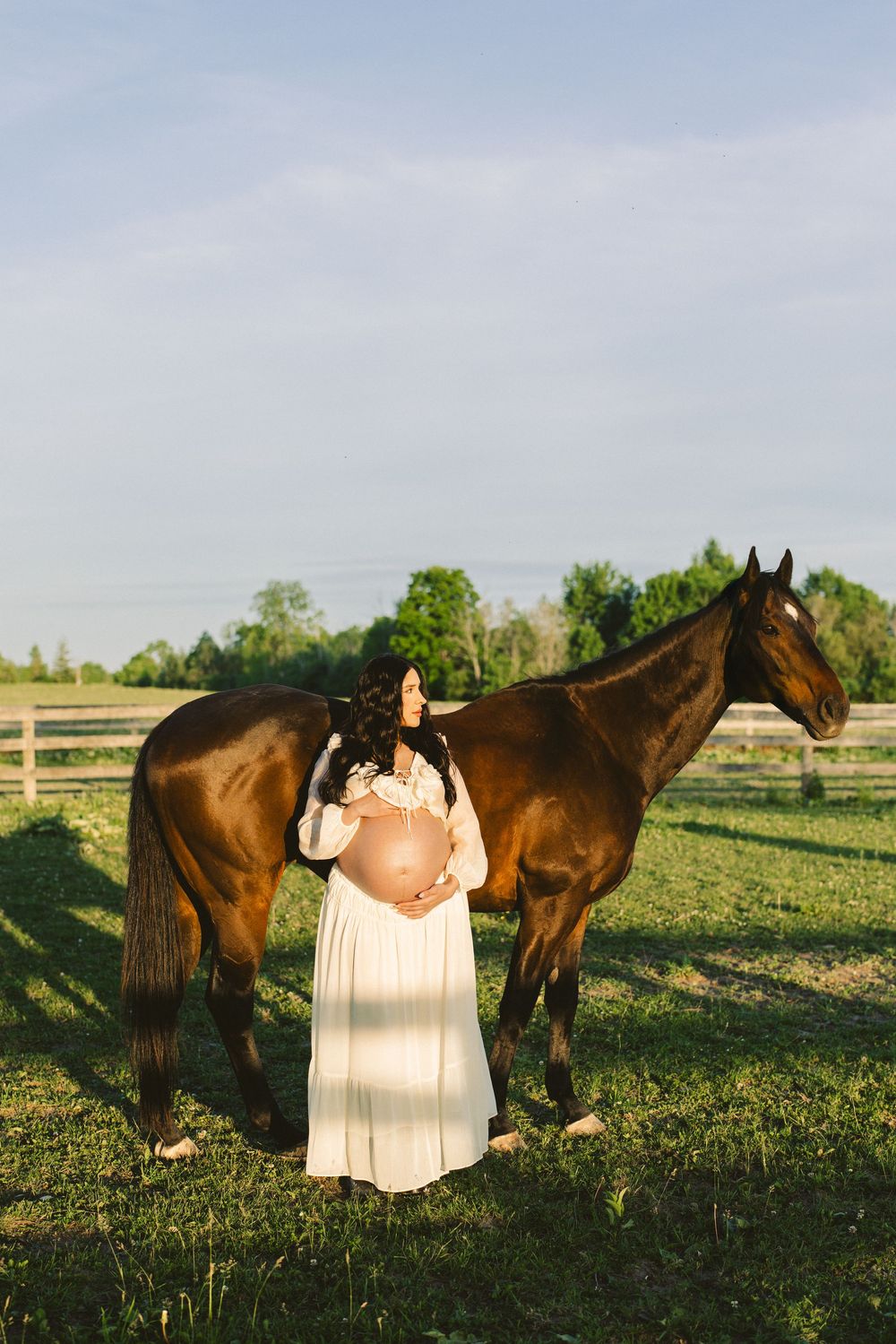 Expectant mother standing between two horses in a green paddock.