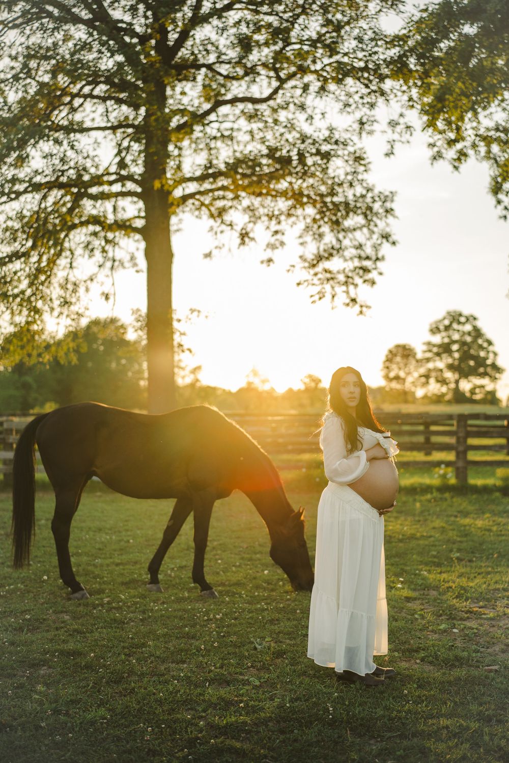 Expectant mother walking through a paddock past a horse at sunset.