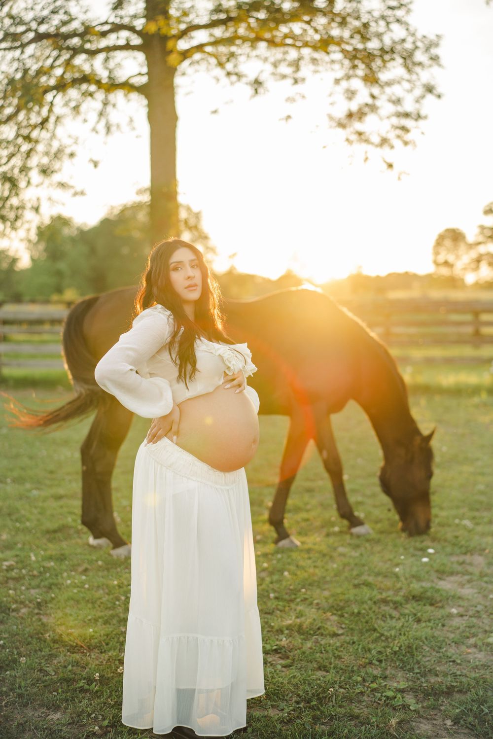 Expectant mother in a white gown beside a grazing horse in warm backlight.