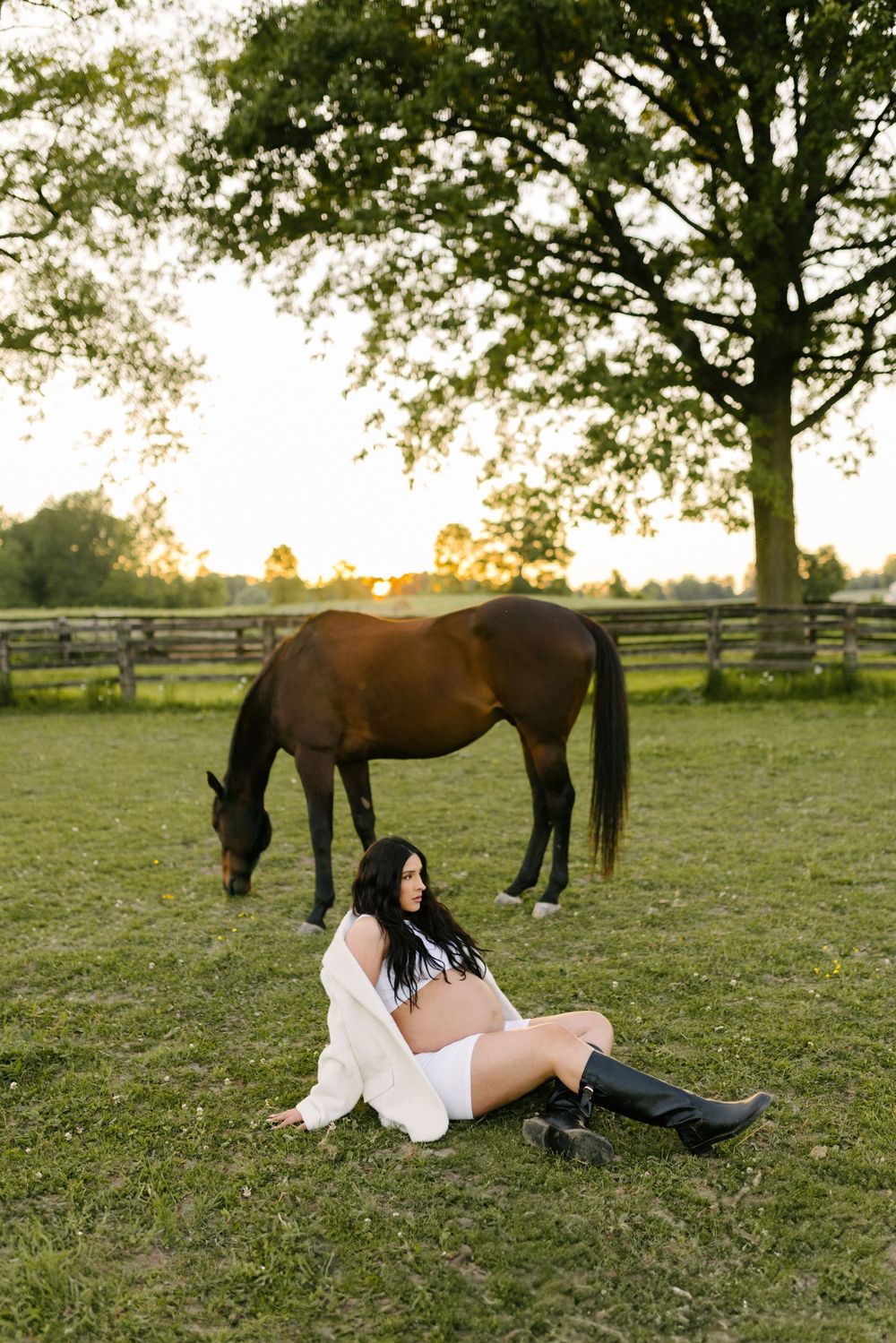 Expectant mother seated on the grass while a horse grazes behind her.