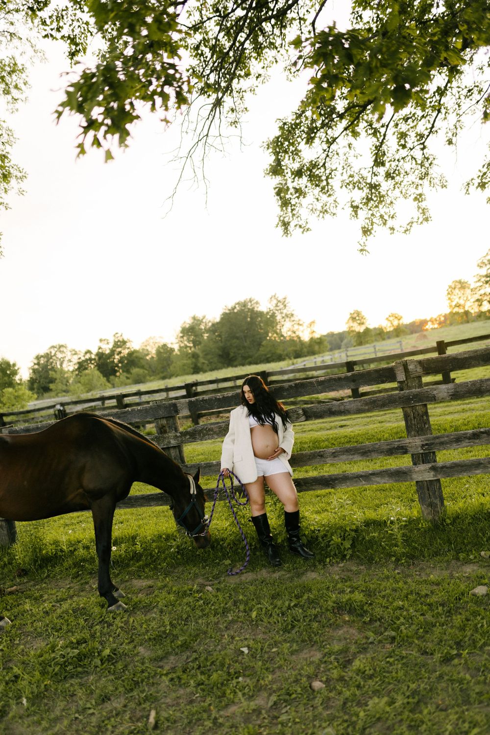 Expectant mother standing by a wooden fence as a horse grazes nearby.