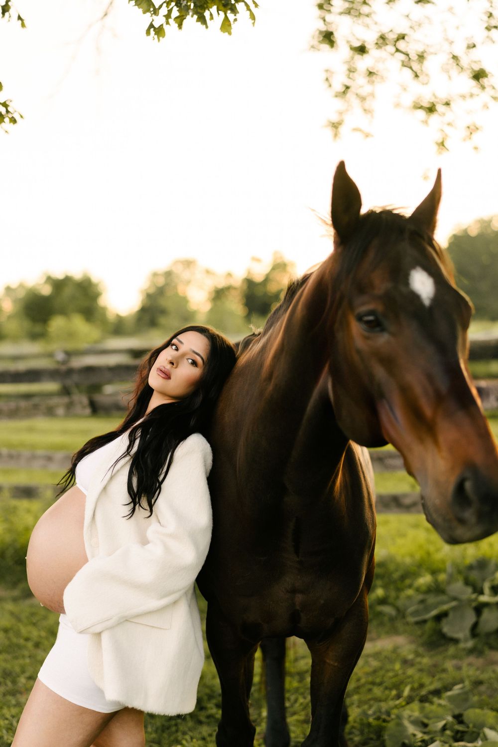 Expectant mother in a knit dress standing beside a brown horse at sunset.