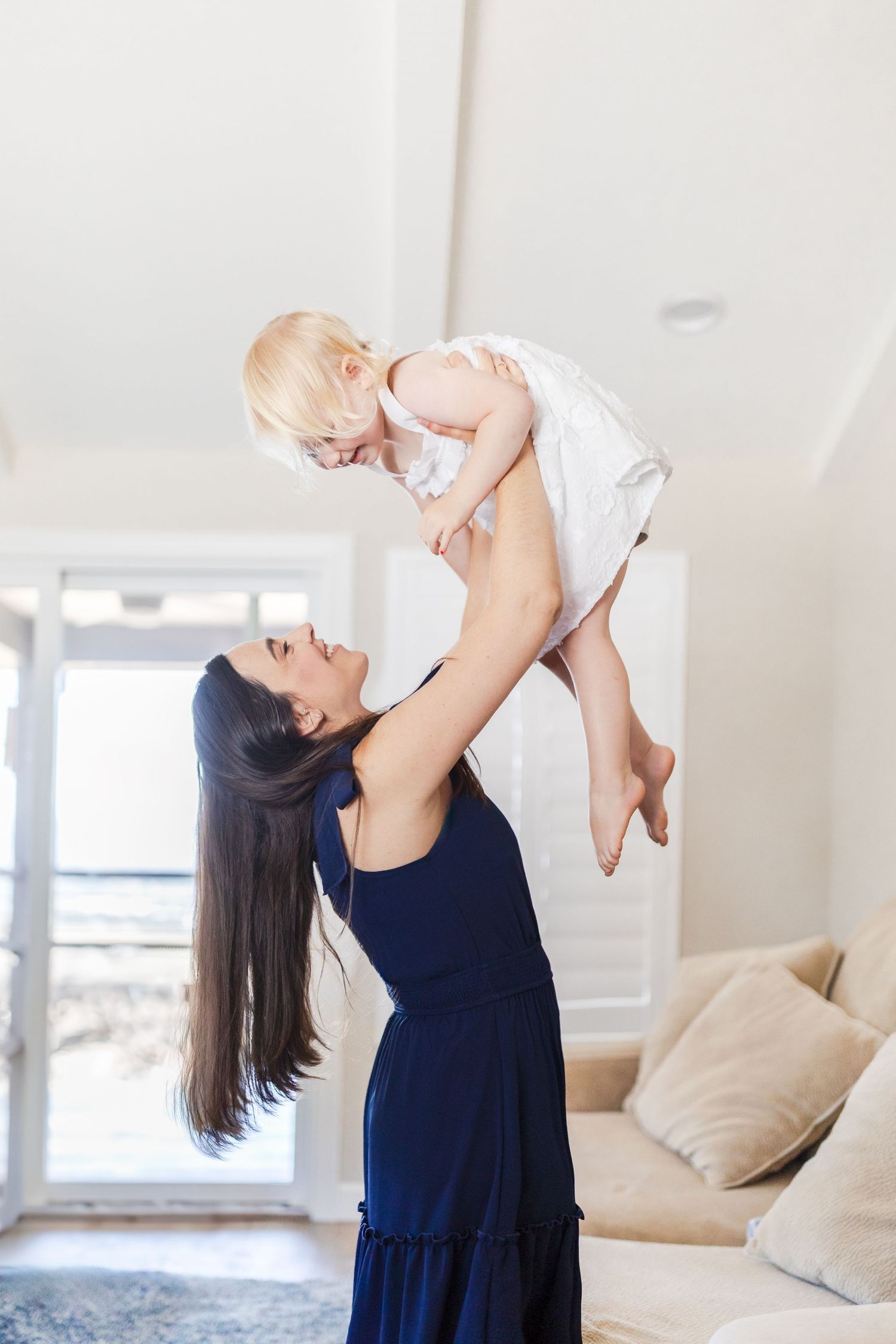Family of four with newborn and toddler smiling on beige sofa with black dog, cozy living room setting.