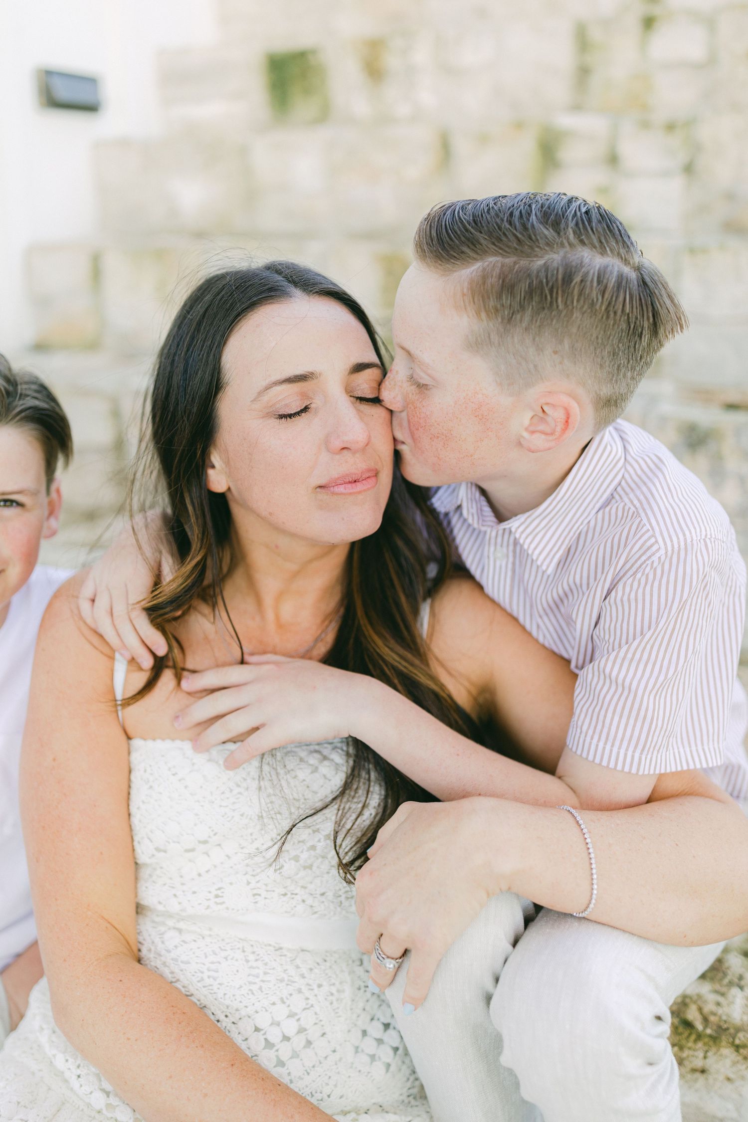 A boy kisses his smiling mother on the cheek during a warm family photo session outdoors.