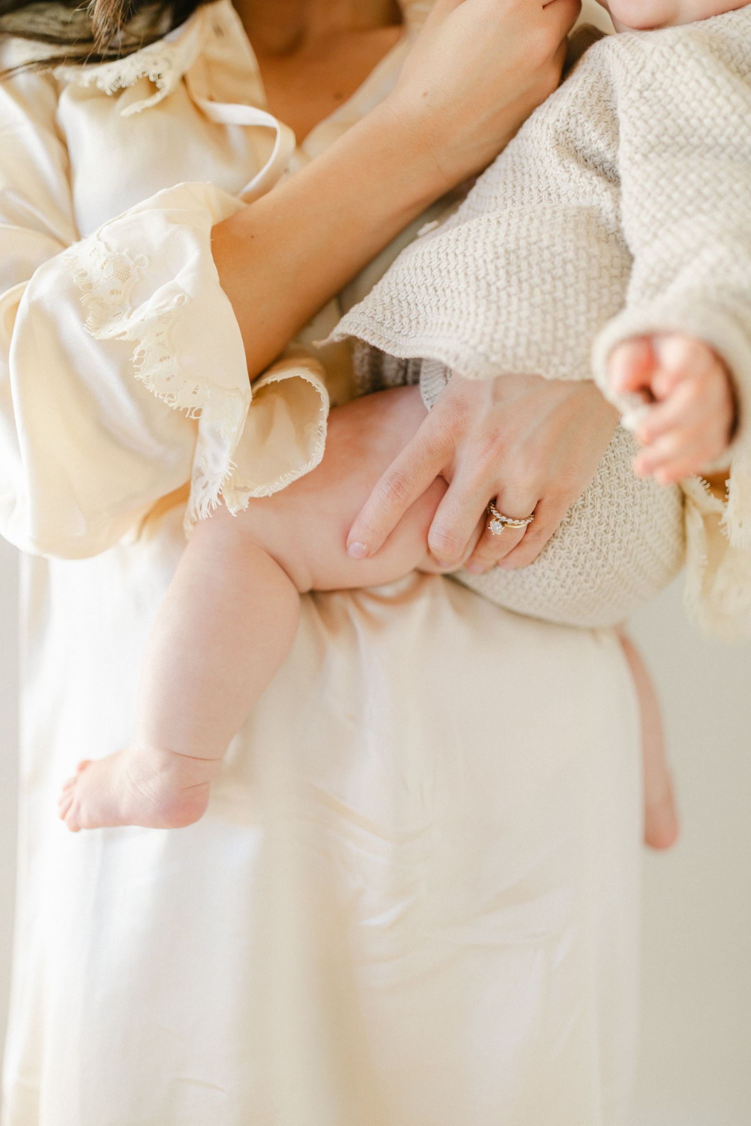 A mother in a cream dress holds her newborn baby, showcasing a delicate ring on her finger.