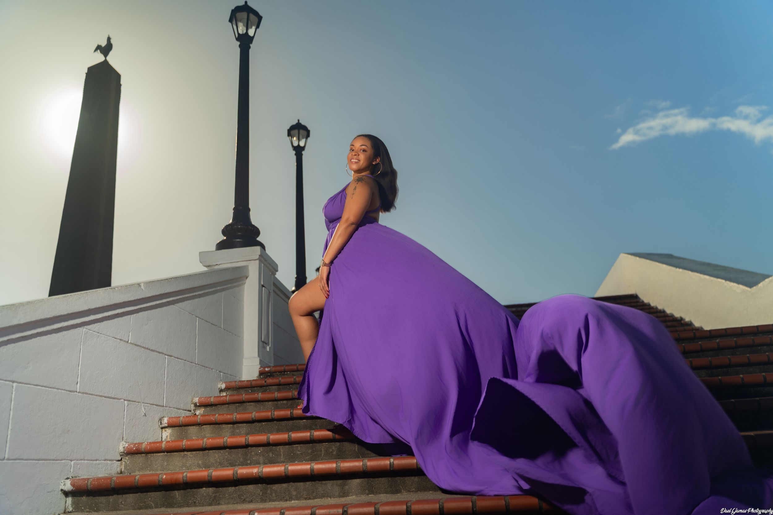 Woman in flowing purple gown poses gracefully on brick steps under bright blue sky with decorative lamp posts.