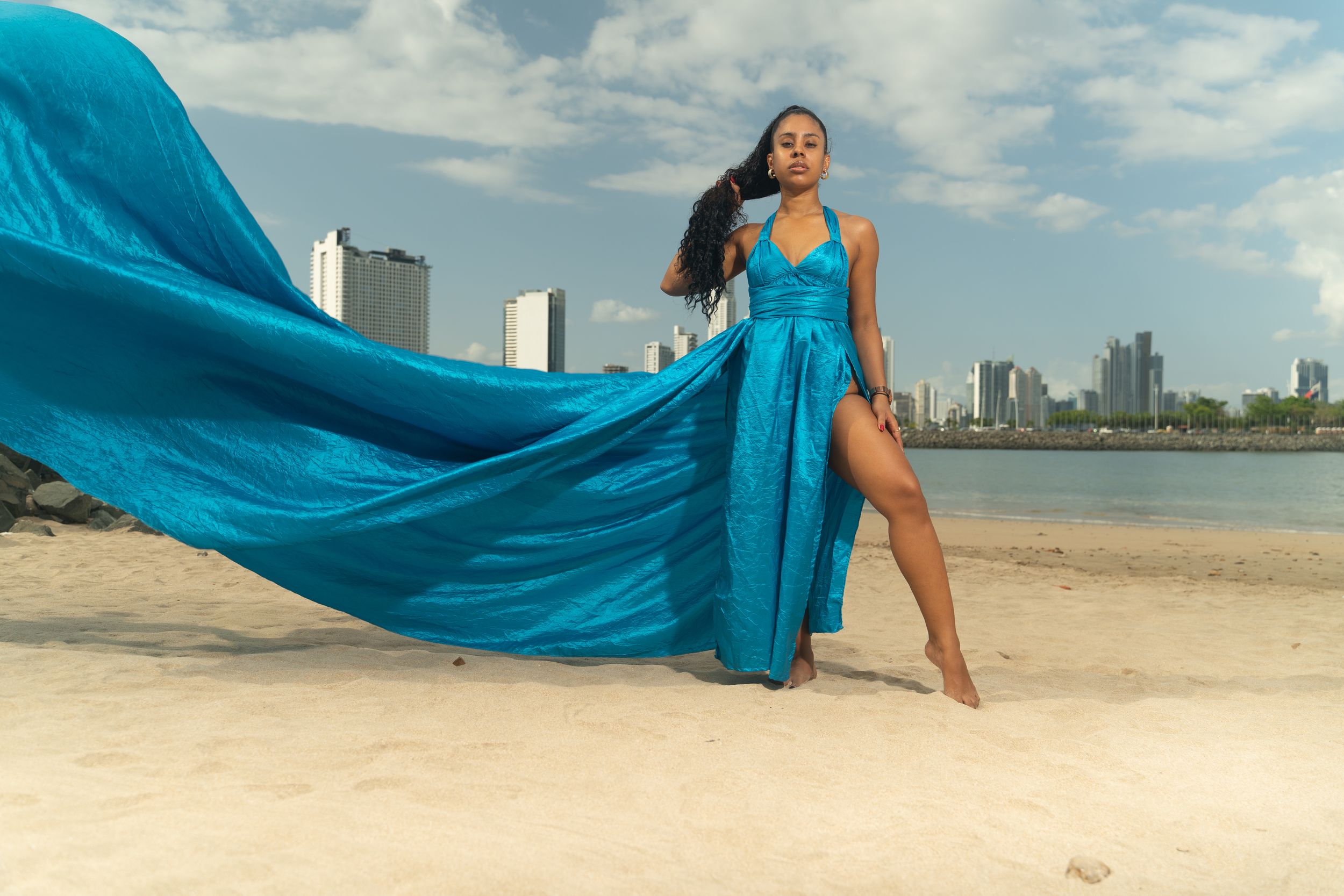 Woman in flowing turquoise gown poses on sandy beach with city skyline backdrop.