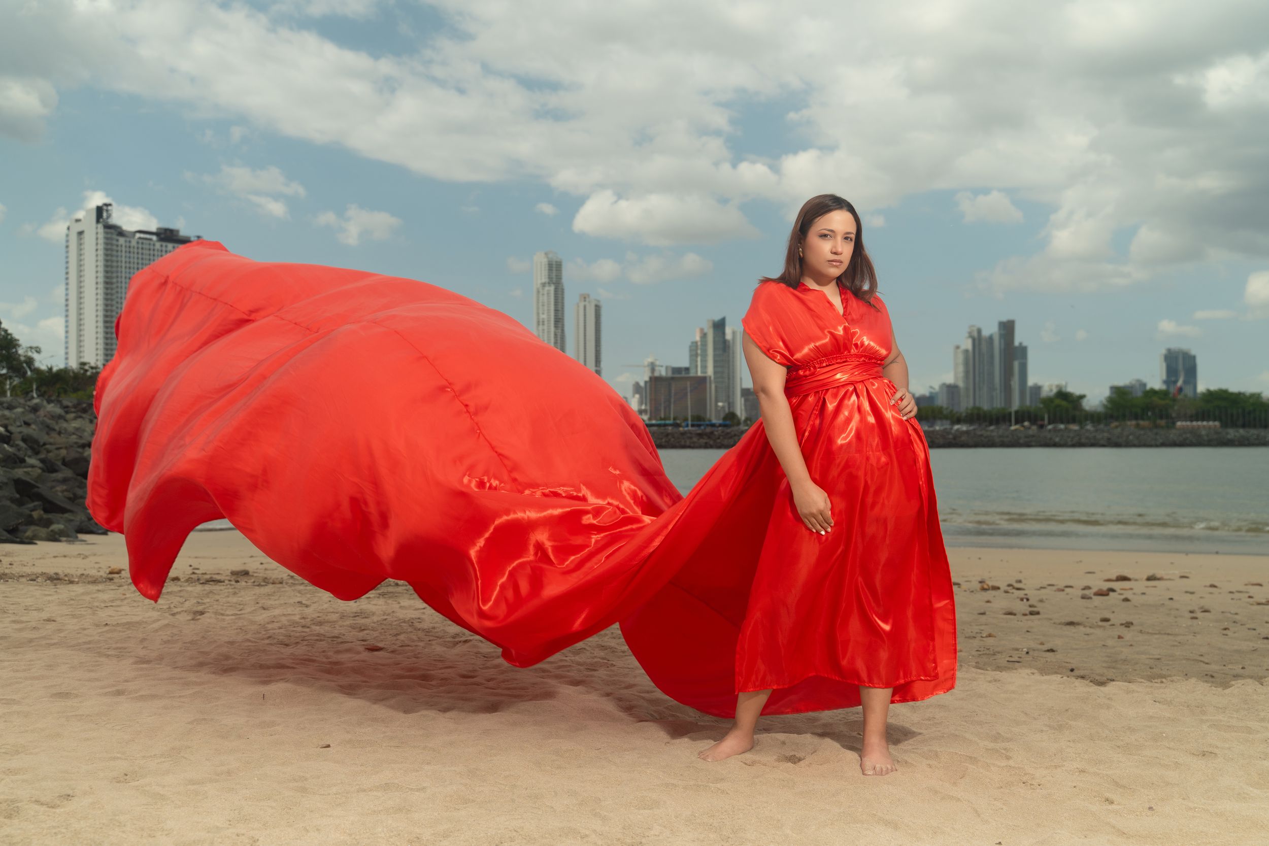 Woman in flowing red dress on beach with city skyline backdrop, fabric billowing dramatically in the wind.