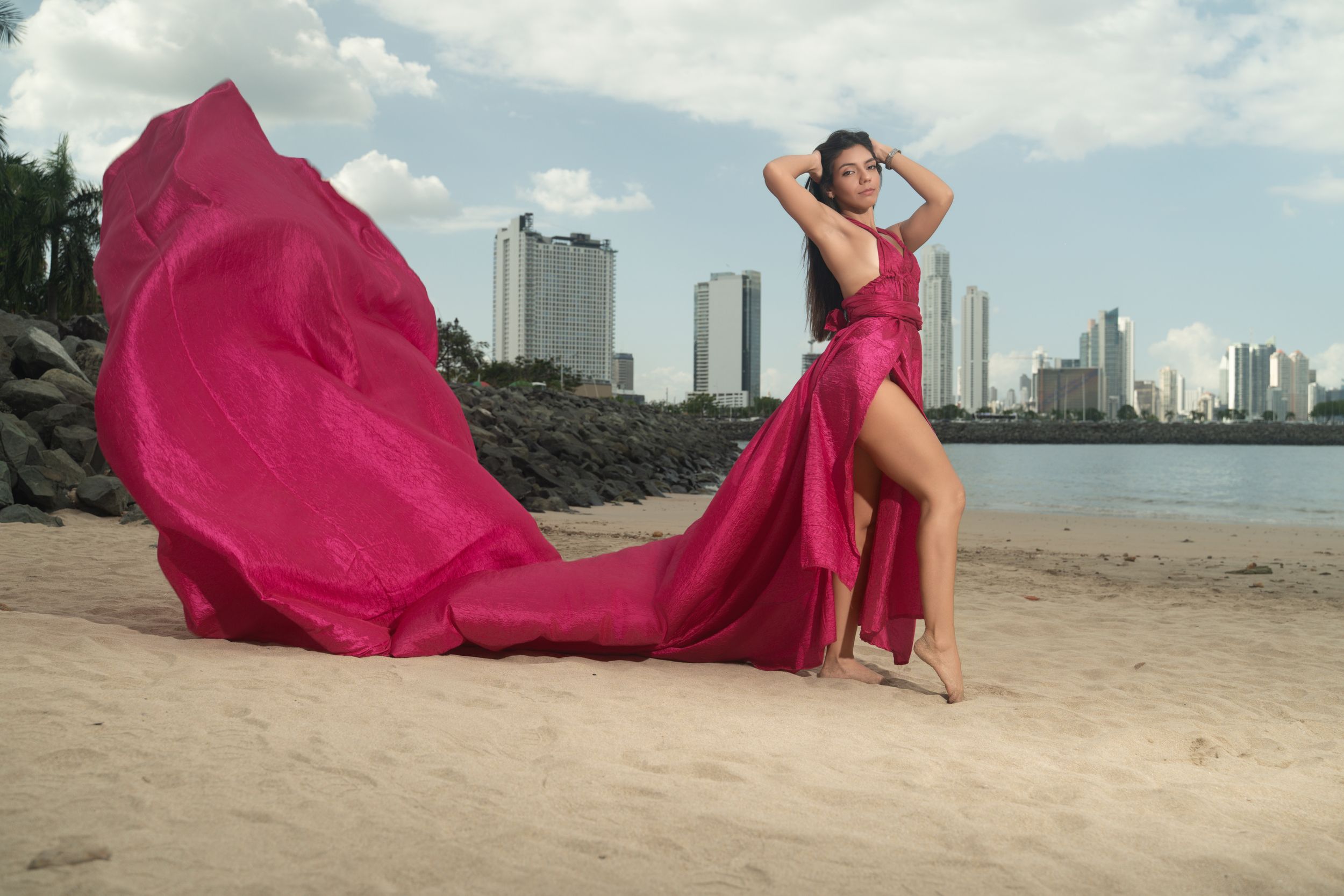 Woman in flowing magenta gown poses on sandy beach with city skyline and water in background.