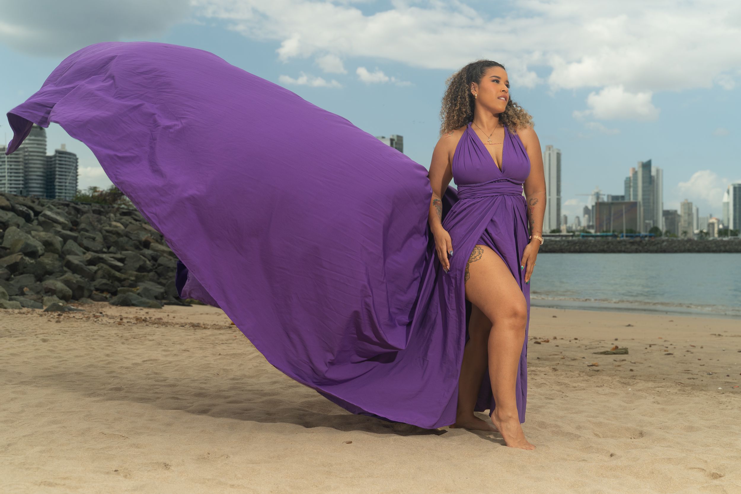 Woman in flowing purple halter dress on sandy beach with city skyline backdrop, fabric billowing dramatically in wind.