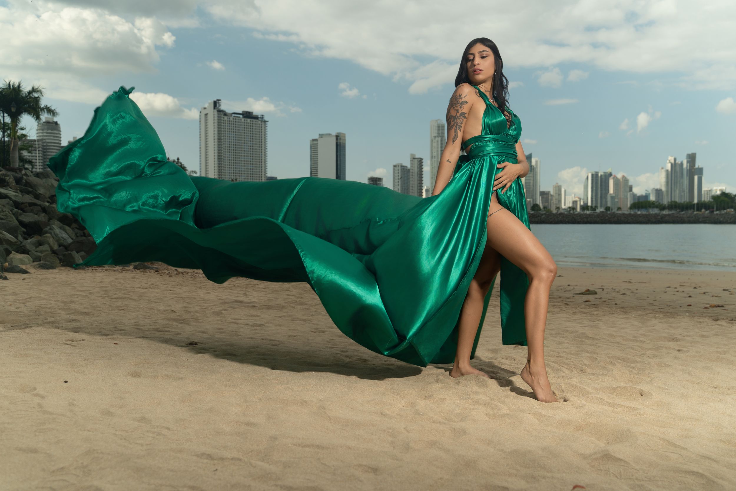 Woman in flowing green satin gown poses on beach with city skyline backdrop, fabric billowing in wind.