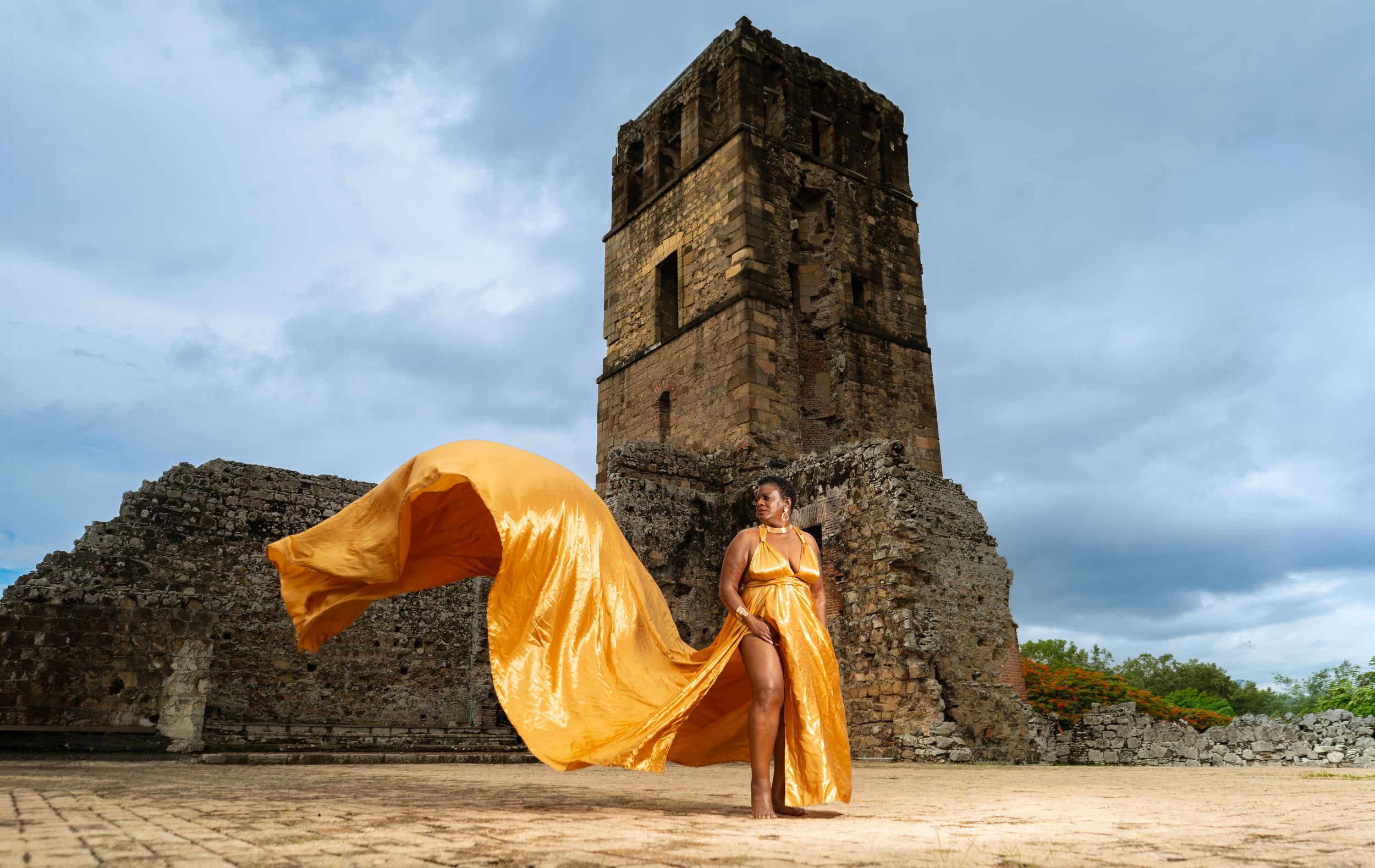 Woman in flowing golden gown poses before ancient stone tower ruins under dramatic cloudy sky.