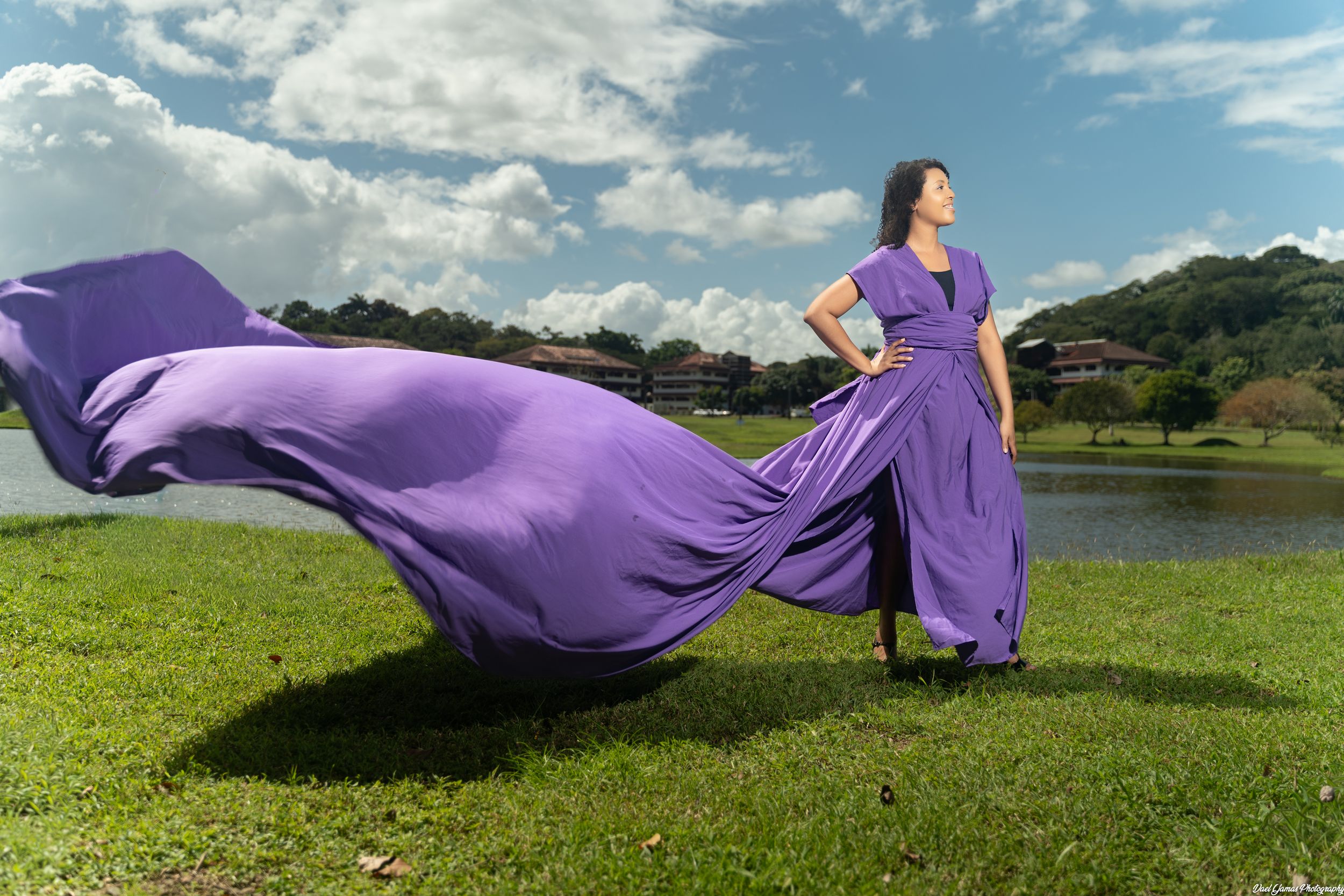 Woman in flowing purple gown standing on green grass, dress billowing in wind against blue cloudy sky near a lake.