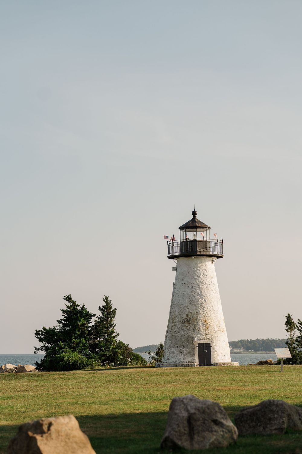 White lighthouse stands on green lawn with trees and water in background under clear sky.