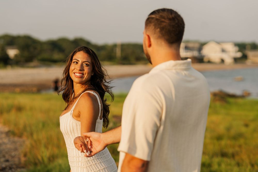 Smiling woman in white dress holds mans hand near scenic coastal marshland at golden hour.