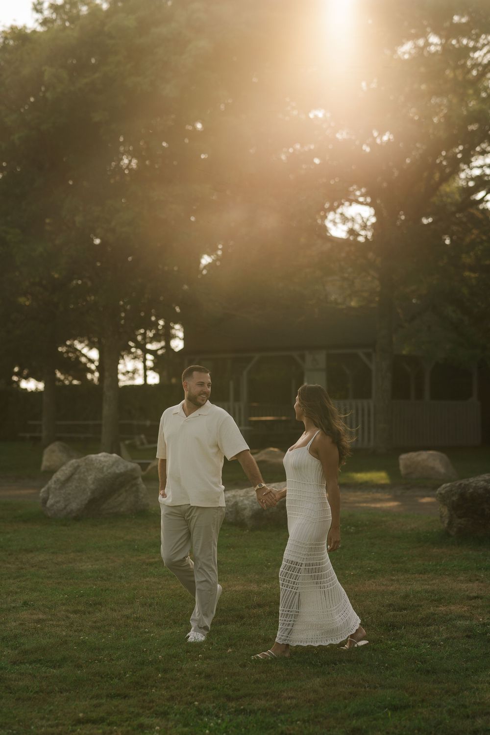 Couple holding hands walking in golden sunset light near gazebo and rocks in a lush green park.