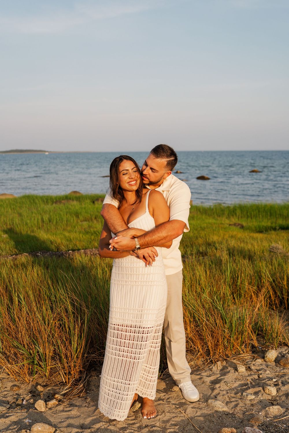 Couple embracing on sandy beach at golden hour, woman in white lace dress, man in white shirt, ocean and marsh grass behind.