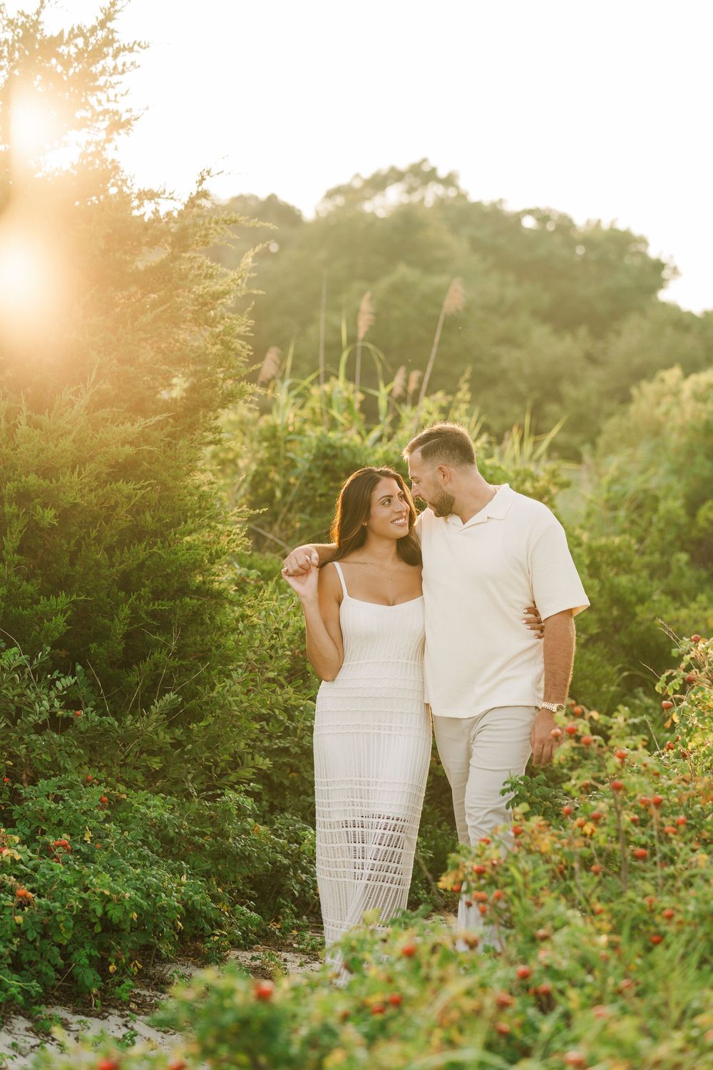 Couple walking through lush green garden at sunset, woman in white dress, man in white shirt, warm golden light.