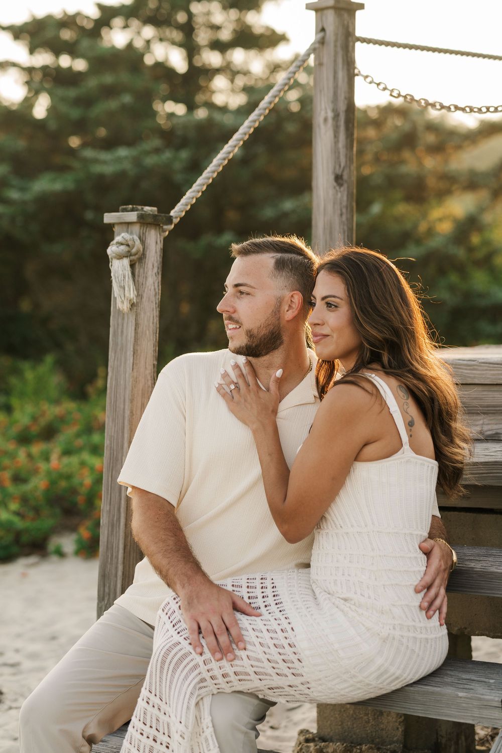 A couple in matching cream outfits sit together on a wooden beach structure during a golden hour photo session.
