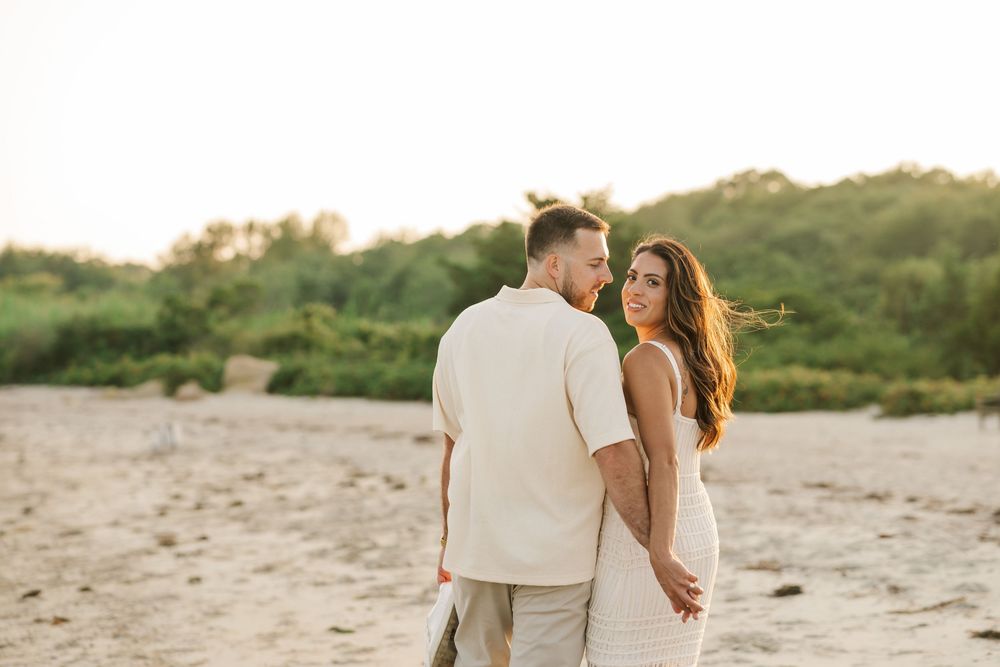 Couple holding hands on sandy beach at golden hour, woman in white dress gazes at man in beige outfit with greenery behind.