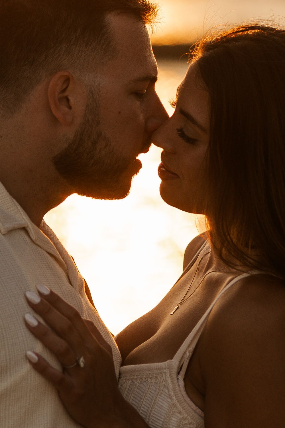 A romantic couple shares a tender kiss at golden sunset, with warm backlit glow highlighting their intimate moment.