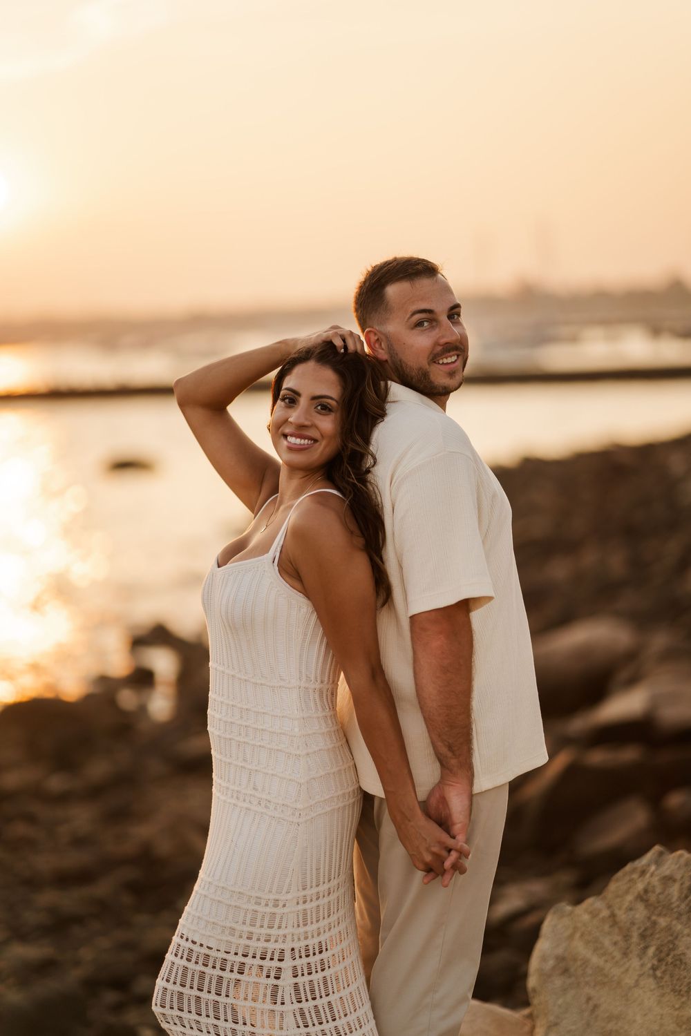 A smiling couple in white outfits hold hands by a rocky shoreline during a stunning sunset engagement shoot.