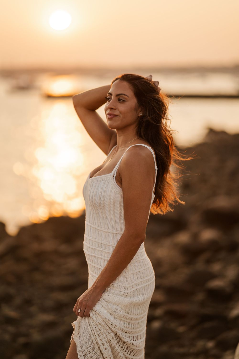 A beautiful woman in a white crochet dress poses gracefully by the ocean during a warm golden sunset.