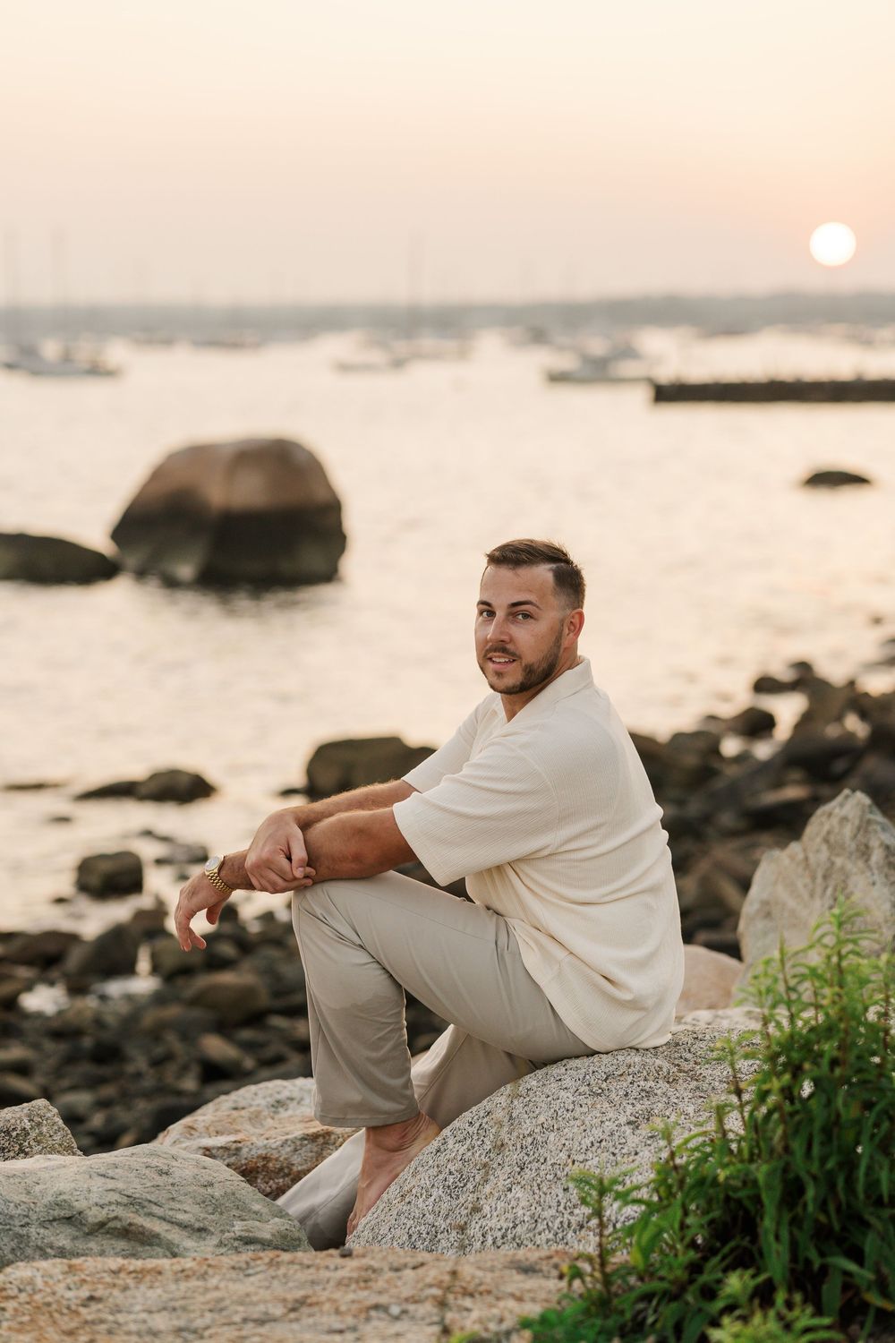 Man sitting on rocky shoreline at sunset with boats and calm harbor waters in background.