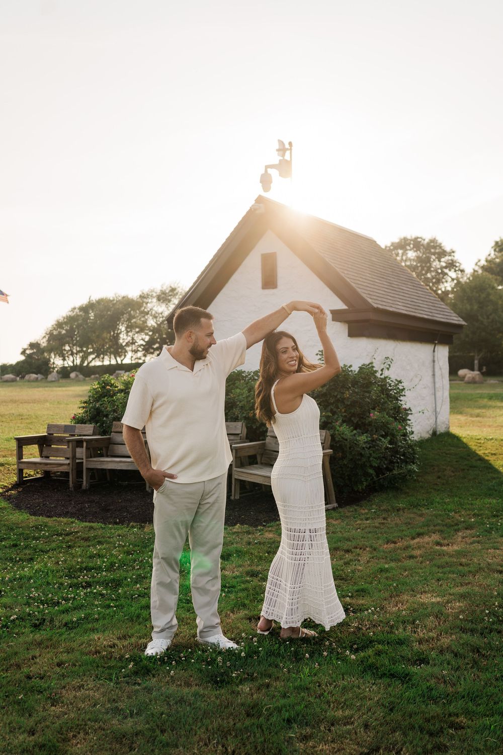 Couple dancing joyfully in front of a white chapel during golden hour sunset on green lawn.
