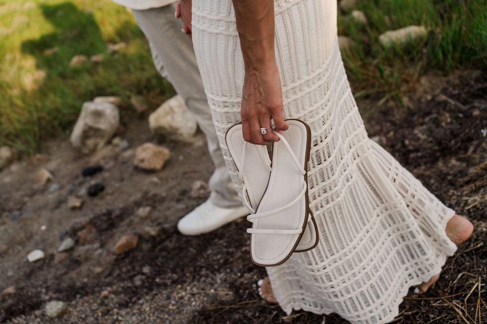 Bride in white lace dress carrying strappy sandals while walking barefoot on a rocky path outdoors.