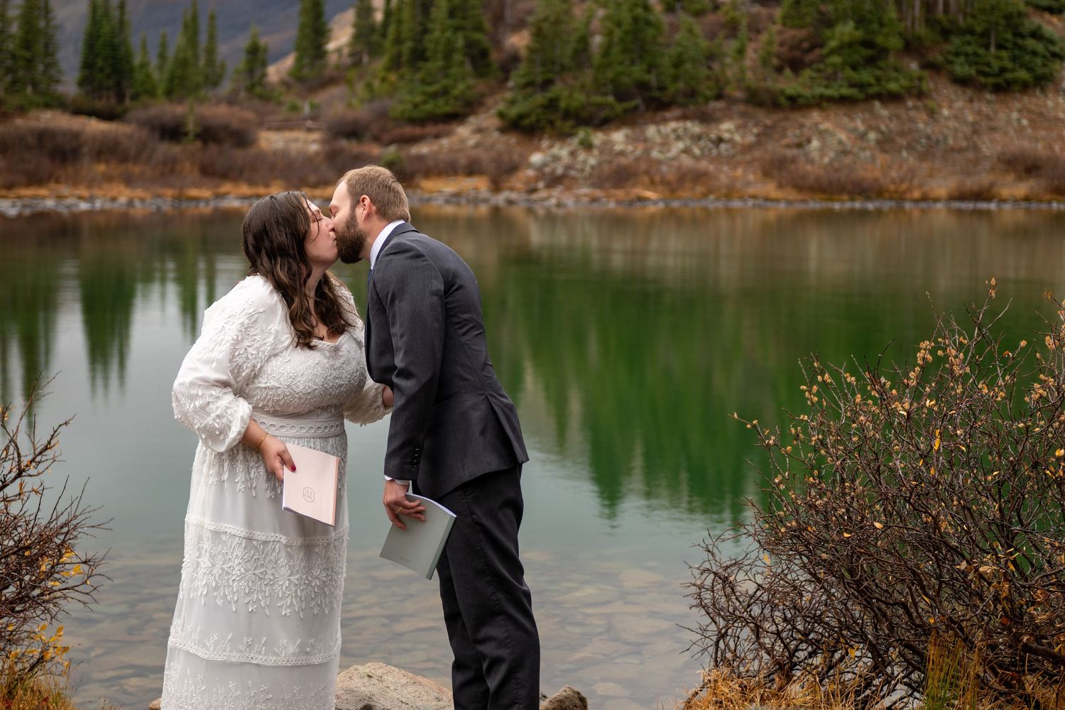 Couple sharing a kiss during outdoor elopement ceremony by a turquoise mountain lake surrounded by evergreen trees.